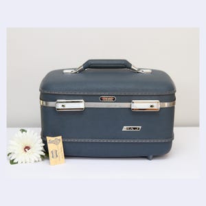 May include: A vintage navy blue vanity case with a silver handle and trim. The case has a rectangular shape with rounded corners and a small tag with text. A white flower is next to the case.