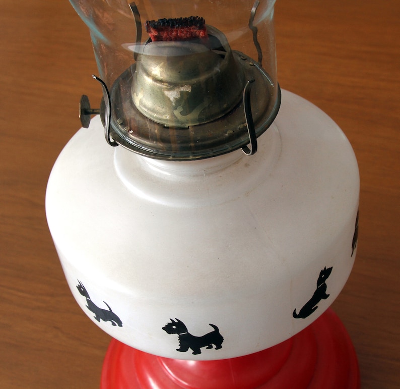 May include: A white oil lamp with a red base and black silhouette images of dogs. The lamp has a clear glass chimney and a brass burner.