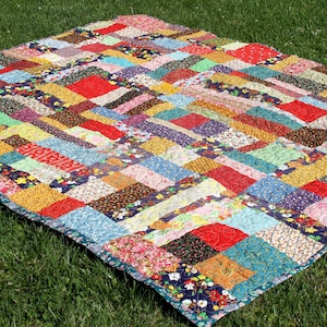 May include: A colorful handmade quilt with a patchwork design. The quilt features a variety of fabric patterns and colors, including red, blue, yellow, and green. The quilt is laid out on a bed of green grass.