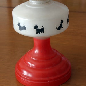 May include: A white oil lamp with a red base and black silhouette images of Scottish Terriers. The lamp has a white frosted glass shade and a red base with a smooth, rounded surface.