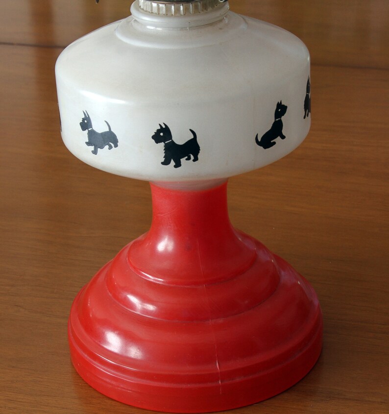 May include: A white oil lamp with a red base and black silhouette images of Scottish Terriers. The lamp has a white frosted glass shade and a red base with a smooth, rounded surface.