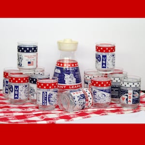 May include: A collection of vintage drinking glasses and a pitcher with patriotic designs. The glasses feature red and blue accents with stars and the text "USA" and "Apollo 12". The pitcher has a white lid.