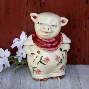 May include: A cream-colored ceramic pig cookie jar featuring a glossy finish, a dark red neck scarf, and painted pink floral accents on the body. The pig has closed eyes with long eyelashes and a smiling expression, with brown painted hooves at the base.