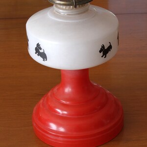 May include: A red and white oil lamp with a white shade featuring black silhouettes of Scottish Terriers. The lamp has a red base with a white shade.