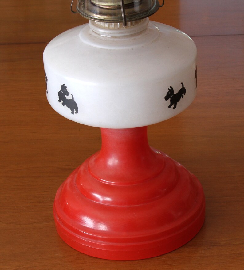 May include: A red and white oil lamp with a white shade featuring black silhouettes of Scottish Terriers. The lamp has a red base with a white shade.