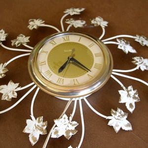 May include: A round wall clock with a gold-toned face, black hands, and Roman numerals. The clock is surrounded by white metal stems extending outward, each tipped with a white and gold leaf-shaped accent. The brand name Rhythm is printed on the face.