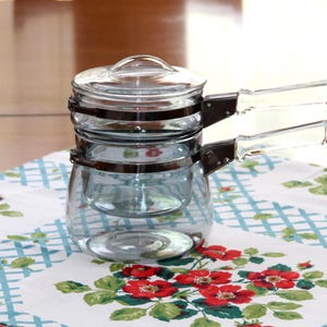 May include: A clear glass double boiler set consisting of two stacked glass pots with a matching glass lid. Each pot features a metallic silver band around the rim and a clear glass handle attached with silver-toned hardware for easy lifting.