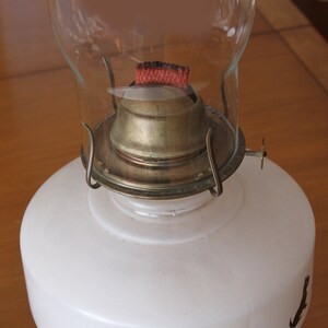 May include: A white glass oil lamp with a brass burner and a red wick. The lamp has a clear glass chimney.