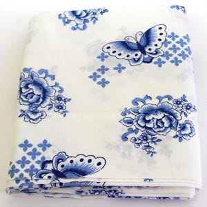 May include: A folded piece of white fabric featuring a repeating pattern of blue butterflies and blue floral clusters. The design includes small four-petaled blue flower accents scattered across the surface, creating a consistent visual motif.