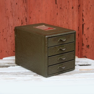 May include: A small, olive green storage cabinet with a textured exterior finish and four stacked drawers. Each drawer features a simple metal pull handle. A rectangular label with red and white text is visible on the top surface of the unit.