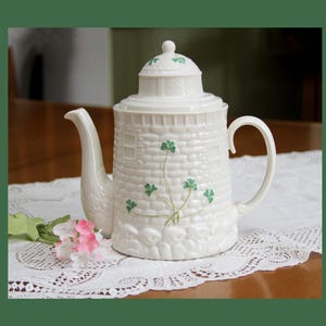 May include: A white ceramic teapot shaped like a lighthouse with a textured brick pattern, a curved handle, and a spout. The body features painted green shamrock motifs, and the lid has a small knob with green shamrock details on top.