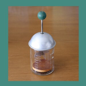 May include: A vintage, silver-colored food mill with a green knob on top. The mill has a glass jar with markings for measuring. A brown food item is inside the jar.