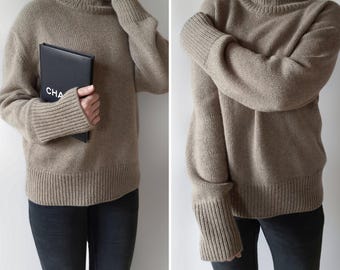 Puede incluir: Un jersey beige de cuello alto con puños y dobladillo acanalados. El jersey se combina con vaqueros negros. Un cuaderno negro con las letras "CHA" se sostiene en la mano izquierda.