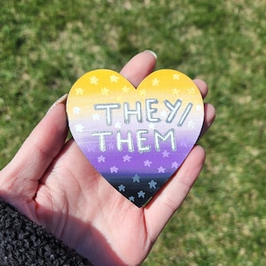 May include: A heart-shaped wooden pin with a yellow, purple, and white gradient. The pin has white stars and the text "THEY/THEM" written in silver glitter.
