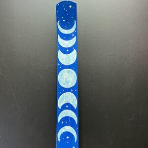 May include: A long, narrow, slightly curved incense holder painted in a deep blue color. The surface features a hand-painted depiction of the lunar cycle, showing various moon phases in light blue, surrounded by small white stars and dots.