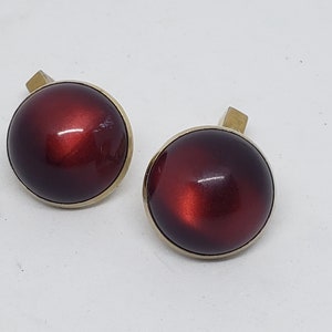 May include: Pair of vintage cufflinks. Each cufflink features a round, glossy, deep red center with a gold-colored border and a gold-colored rectangular post. The cufflinks are set against a white background.