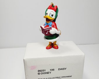 May include: A Christmas ornament featuring Daisy Duck, a Disney character, dressed in a red and green holiday outfit, holding a book titled "CAROLS". The ornament is on a white box with the text "26231 106 DAISY ©DISNEY THIS IS A COLLECTIBLE ORNAMENT AND NOT A CHILD'S TOY"