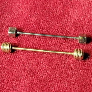May include: Two collar bars, one silver-toned with smooth square ends and one gold-toned with ribbed cylindrical ends. Each bar features a thin metal rod connecting two screw-on ends, designed to be worn across the collar of a dress shirt.