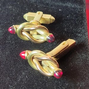 May include: A pair of gold-toned cufflinks with a knot design. Each cufflink features a knot with a red gemstone at each end. The cufflinks are displayed on a black surface.