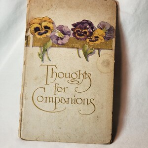 May include: Antique book titled "Thoughts for Companions." The cover features a floral design of yellow, purple, and white pansies above the title in gold script. The book has a worn, aged appearance.