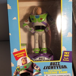 May include: A plastic Buzz Lightyear toy bank with a purple base and a green and white space ranger suit. The toy has a button on the base that says "Try Me! Push Button". The box also says "Original Voice" and "Buzz Lightyear Electronic Talking Bank".