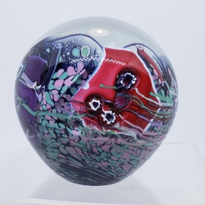 May include: A glass paperweight with a red, purple, green, and white abstract design. The paperweight is round and has a clear glass base.
