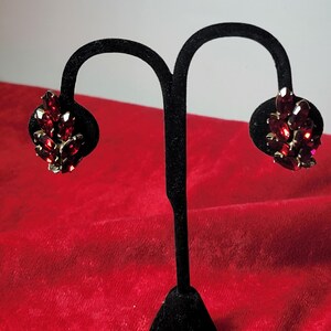 May include: A pair of red rhinestone earrings on a black earring stand. The earrings are made of a cluster of red rhinestones.