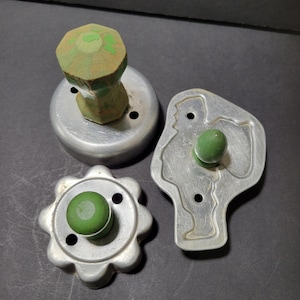 May include: Three vintage cookie cutters. One is round with a wooden handle, another is a flower shape, and the third is a person-shaped cutter. All are made of metal and have green handles.