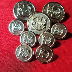 May include: A collection of silver-colored buttons with the text "Santa Fe" in a square design. The buttons are arranged on a red surface. The buttons vary in size and shape, with a mix of round and oval designs.