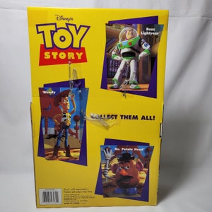 Peut inclure: Afficheur en carton pour les personnages de Toy Story repr&eacute;sentant Woody, Buzz l'&Eacute;clair et M. Patate. L'afficheur est jaune avec des images des personnages et le texte "Disney's Toy Story" et "Collect Them All!"
