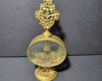 May include: A gold-toned perfume bottle with a decorative floral design on the stopper and a clear glass body. The bottle has a round base and a detailed floral design on the bottom.