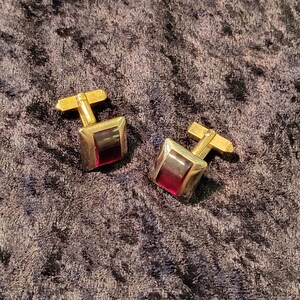 May include: A pair of gold cufflinks with red glass insets. The cufflinks are square shaped and have a simple design.