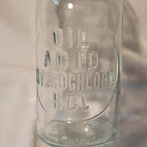 May include: A clear glass apothecary bottle with a wide neck and a rounded body. Embossed on the front is the text DIL ACID HYDROCHLORIC HCL within a circular outline. The base features additional embossed text, including the word POTASH.