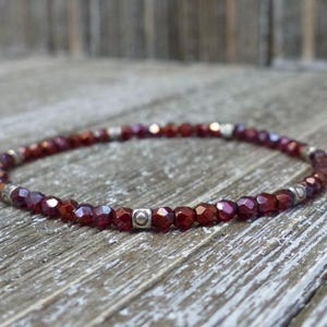 May include: A beaded bracelet featuring faceted, deep red beads and small silver-colored spacers. The bracelet is strung on an elastic cord and rests on a weathered wooden surface. The bracelet is a piece of jewelry.