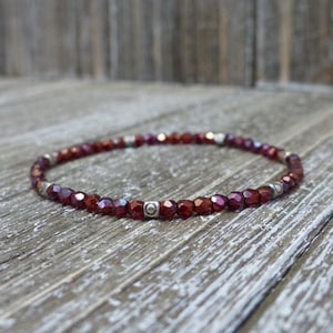 Puede incluir: Una pulsera de cuentas con pequeñas cuentas facetadas de color rojo intenso. La pulsera tiene espaciadores plateados y se muestra sobre una superficie de madera envejecida. Las cuentas tienen un aspecto brillante, similar a una gema.