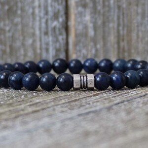 May include: A dark blue beaded bracelet with a silver clasp. The bracelet is made of round, polished beads. The bracelet is resting on a weathered wooden surface.