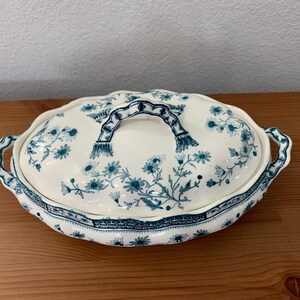 May include: A white oval serving dish with a blue floral pattern and a lid with a blue handle.