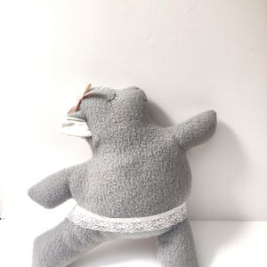 May include: A gray plush toy with a white lace trim and a pink bow on its head. The toy is in a yoga pose with one leg raised.