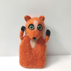 May include: A small orange felt fox with black arms and green eyes. The fox is standing on a white background.