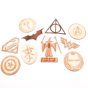 May include: A set of 10 wooden coasters featuring various pop culture icons, including Batman, Superman, Wonder Woman, Captain America, Iron Man, Thor, Gandalf, the One Ring, the Deathly Hallows symbol, and the Avengers logo. The coasters are made of natural wood and have a rustic, hand-crafted look.