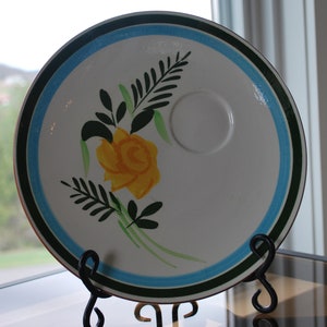 May include: A white ceramic plate with a blue and green border. The plate features a yellow rose with green leaves and a small, white, circular indentation in the center.
