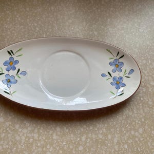 May include: Oval-shaped white ceramic dish with a brown rim and a central recessed area. The dish is decorated with blue floral designs on each end, featuring blue flowers and green leaves. A decorative serving piece.