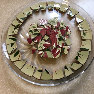 May include: A clear glass plate with a floral design, holding broken pieces of a ceramic plate. The ceramic pieces have a light green background with red and white floral patterns and green leaves. The broken pieces are arranged in a circular pattern.