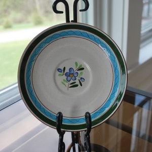 May include: A white ceramic saucer with a blue and green floral design. The saucer is on a black metal stand.