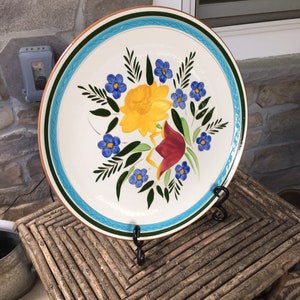 May include: A large, round, white ceramic plate with a blue, green, and orange border. The plate features a hand-painted floral design with yellow, red, and blue flowers.