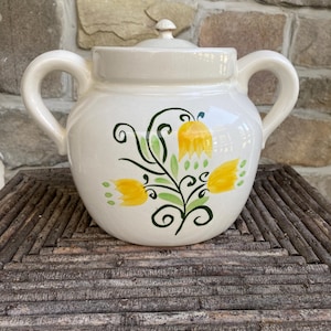 May include: Cream-colored ceramic pot with a lid and two handles. The pot is decorated with a floral design featuring yellow tulips and green leaves. The pot is sitting on a wooden surface.