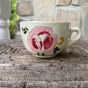 May include: Cream-colored ceramic teacup with a large pink and burgundy floral design. The cup has a rounded shape, a small handle, and a yellow interior rim. The cup is placed on a weathered wooden surface.