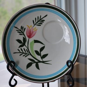 May include: White ceramic plate with a floral design featuring a pink tulip and green leaves. The plate has a blue and green border and a recessed center for a cup. It is displayed on a black metal stand.