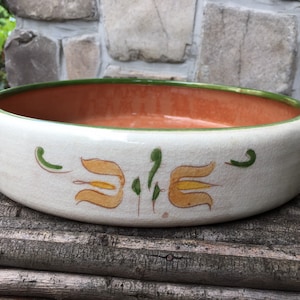 May include: A white ceramic bowl with a green rim and orange interior. The bowl has a hand-painted design of stylized yellow flowers with green leaves.
