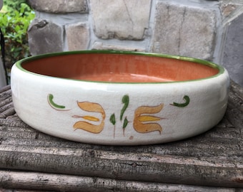 May include: A white ceramic bowl with a green rim and orange interior. The bowl has a hand-painted design of stylized yellow flowers with green leaves.